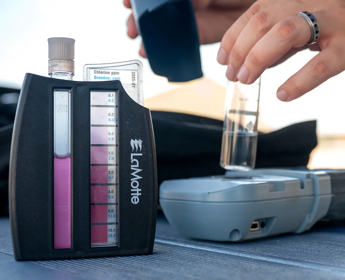 A hand filling a sample tube beside a chlorine and pH comparator block on a pool deck.